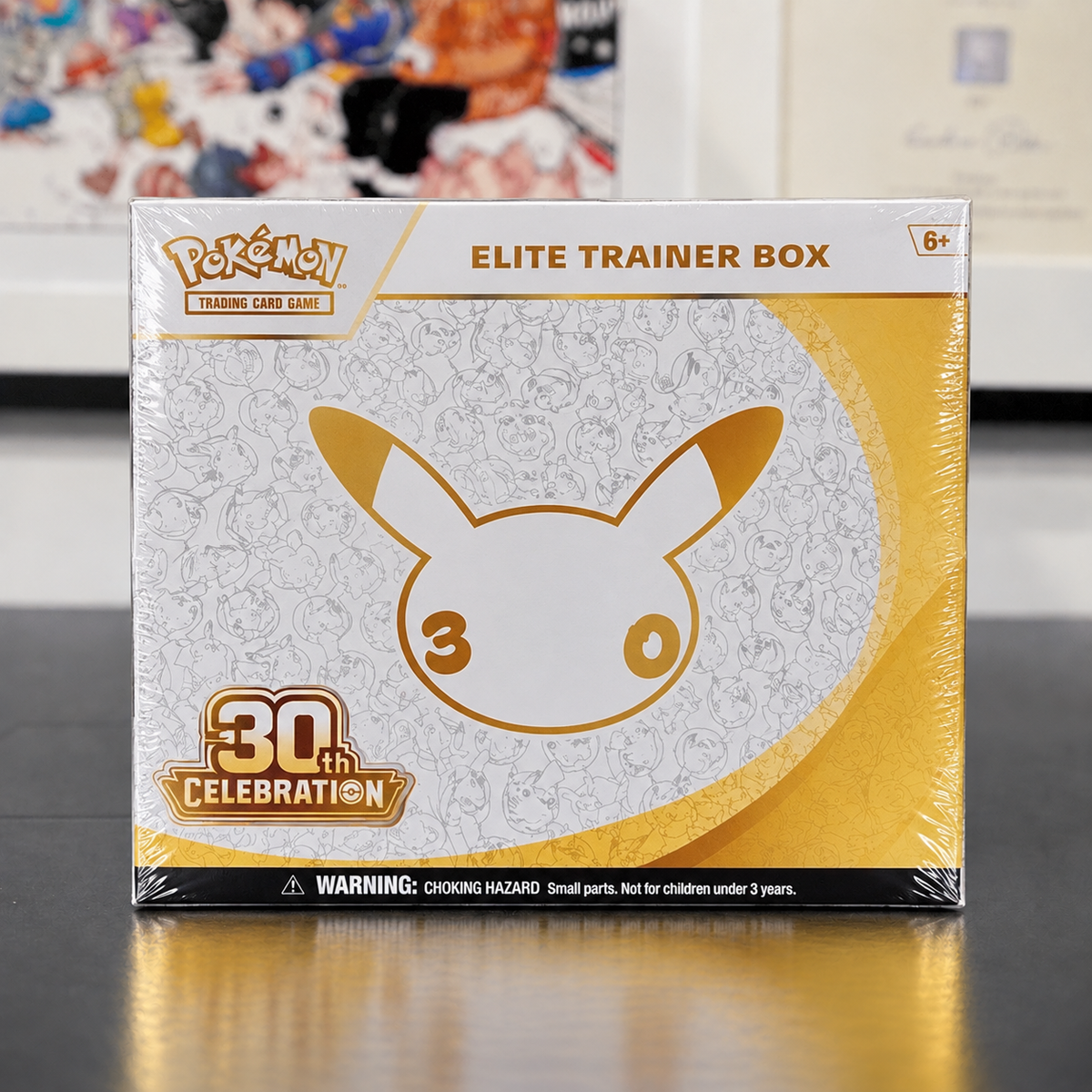 Pokémon Elite Trainer Box 30th Celebration sealed in plastic, featuring a white and gold Pikachu design on the front, set on a reflective dark surface with a blurred background.
